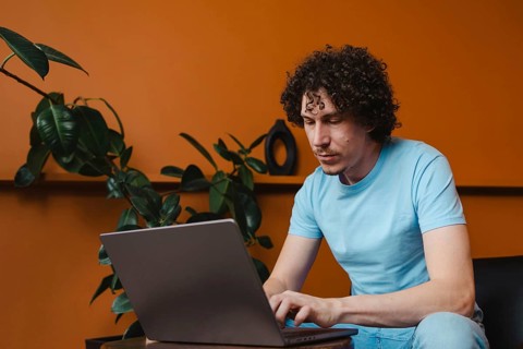 A young man with curly hair focused on his laptop in a modern room with orange walls and green