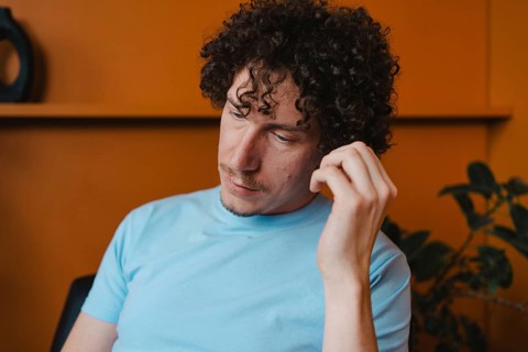 A young man with curly hair in a light blue shirt, sitting thoughtfully in a room with an orange