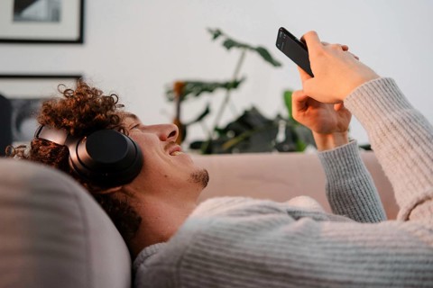 A young man with curly hair enjoying leisure time on a sofa, wearing headphones and using a