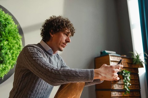 A young man with curly hair uses a smartphone, sitting beside a vibrant green wall. This scene