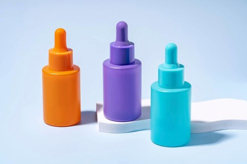 Three serum bottles in vibrant orange, purple, and turquoise colors, showcased against a soft blue
