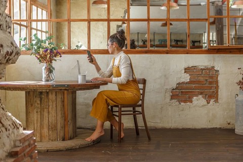 A young woman uses a smartphone and laptop while sitting at a round wooden table in a rustic cafe,