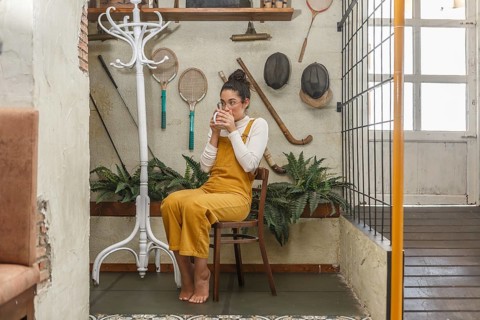 A woman in a yellow jumpsuit sips coffee in her eclectic teleworking space, surrounded by vintage