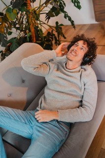 Young man with curly hair relaxes on a modern gray couch, wearing headphones and enjoying music. He