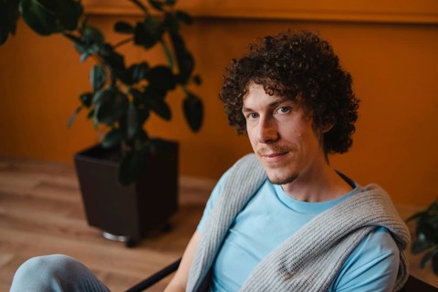 Young man with curly hair in a cozy setting, wearing a blue shirt and a knit sweater. Warm-toned