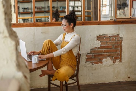 A young woman in a stylishly rustic space focuses on her laptop screen while teleworking, her