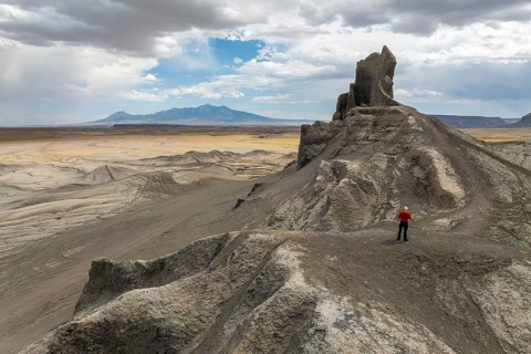 A person in a red jacket stands atop a rugged, rocky outcrop in a vast desert landscape in