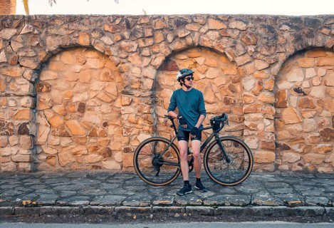 A young male cyclist stands next to his road bike, leaning against an aged stone wall in Zahara de