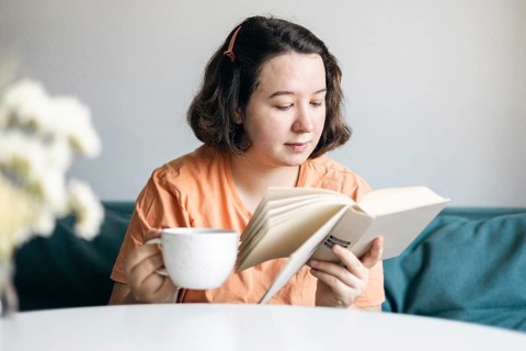 Young woman reads intently while holding a cup of coffee, comfortably seated on a sofa at home,