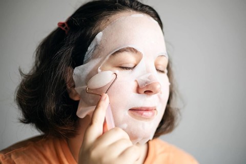 Image of a woman at home, using a facial pink quartz roller while wearing a sheet mask to enhance