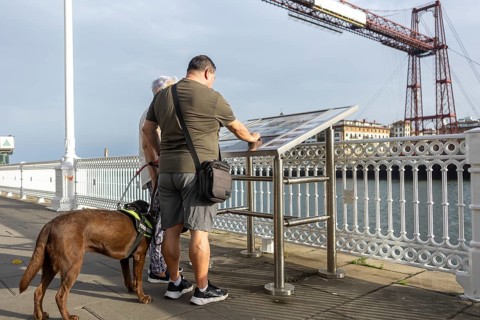 A blind man with a guide dog read an information board on a bridge overlooking a scenic river and