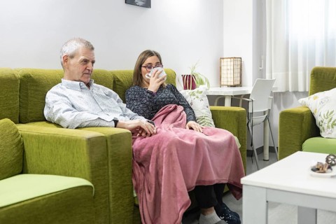 A couple with intellectual disabilities sit comfortably on a green sofa wrapped in a pink blanket.