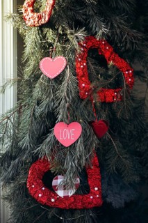 Valentine's Day ornaments in heart shapes hang from evergreen branches, featuring red and pink hues