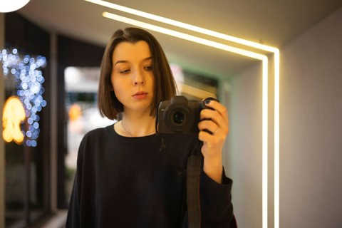 A woman capturing a self-portrait using a DSLR camera in a well-lit interior with neon accents and