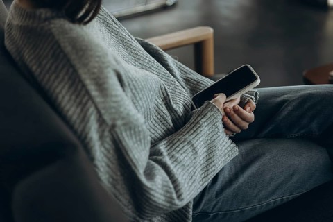 A person wearing a comfy sweater and jeans relaxes on a sofa, holding a smartphone. The setting