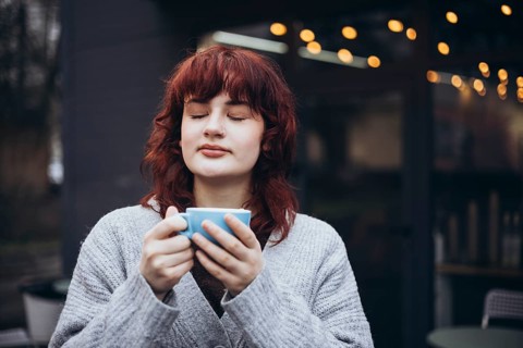A young woman with red hair, in a gray sweater, enjoys a coffee on cafe terrace. String lights