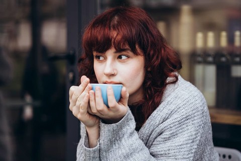 A young woman with red hair sips from a blue cup of coffee, wrapped in a cozy sweater. She gazes
