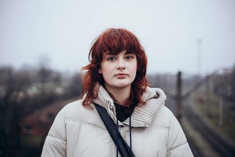 A young woman with red hair stands outdoors wearing a beige winter coat. The background is blurred
