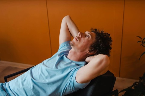 A curly-haired man leans back in a chair with his hands behind his head, gazing upward. The warm