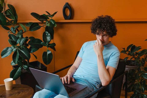 A curly-haired young man in a light blue shirt works on his laptop in a stylish room with plants