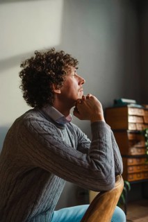 A contemplative man with curly hair sits by a window, resting his chin on his hands. Warm natural
