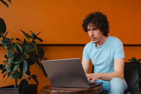 A curly-haired young man focused on his laptop, sitting in a modern space with orange walls and