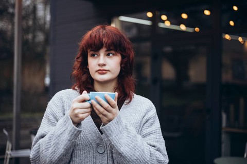 A young woman with red hair, wearing a gray cardigan, holding a coffee cup and gazing thoughtfully