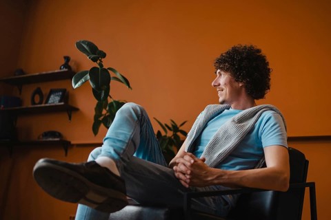 A curly-haired young man sits comfortably in a modern living room with a warm orange background,