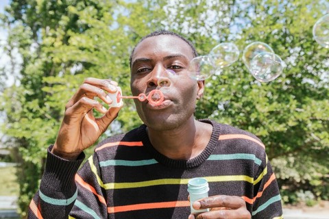 A man in a striped sweater joyfully blows bubbles outside on a bright sunny day, surrounded by lush