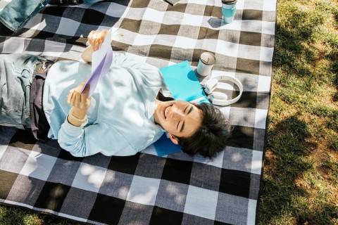 Friends lies on a checkered picnic blanket, enjoying a sunny day with headphones, papers, and