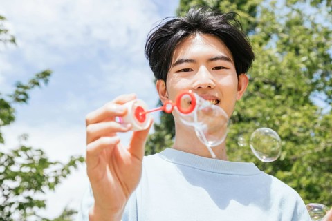 A smiling man enjoys blowing soap bubbles in a sunlit park. The clear sky and vibrant greenery