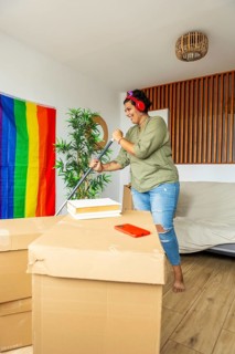 A trans woman smiling while organizing her new home. Surrounded by moving boxes and a pride flag,