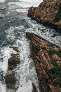 A dramatic aerial view of a rocky coastline where powerful ocean waves crash against rugged cliffs.
