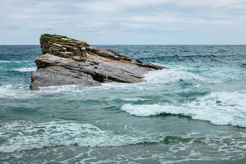 A scenic view of a rocky cliff surrounded by ocean waves crashing under a cloudy sky. The intricate