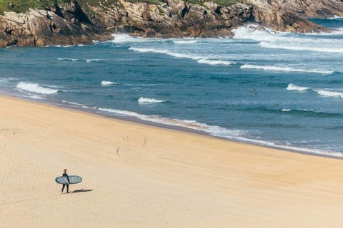A surfer carrying a surfboard walks alone on a sandy beach toward the ocean, with waves crashing