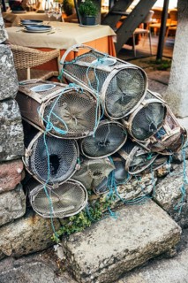 A stack of fishing traps rests against a rough stone wall beside an outdoor cafe table. The scene