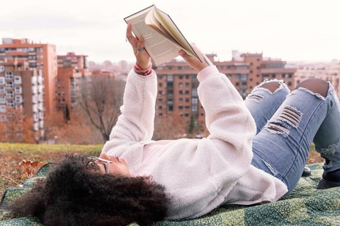 A woman lies on a blanket in a park, reading a book. They wear casual clothes, including jeans and