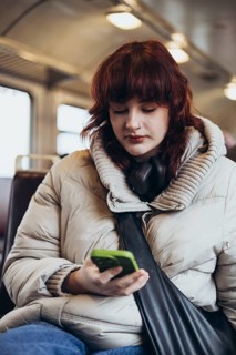 A young woman in a cozy coat, with headphones around her neck, focuses on her smartphone while