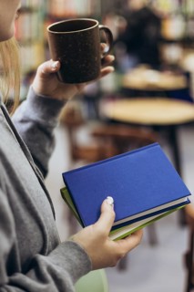A woman in casual attire holds a coffee mug and books, enjoying a quiet moment in a cozy bookstore