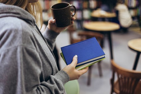 A woman in a gray hoodie holds books and a cup of coffee, enjoying a relaxed moment in a cozy