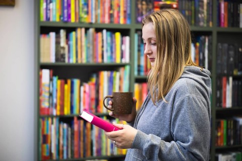 A woman in a gray hoodie enjoys a quiet moment in a bookstore. They hold a pink book and a brown