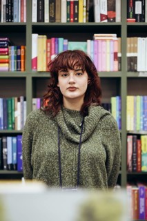 A young female academic pauses thoughtfully in a library, surrounded by colorful bookshelves filled