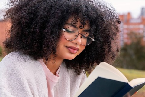 A young latin woman with curly hair and glasses reads a book outdoors, enjoying a peaceful and
