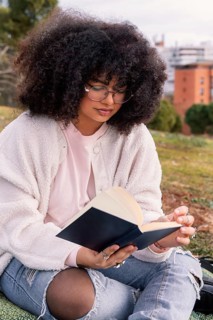 A latin woman with curly hair and glasses enjoys reading a book while seated on grass in a peaceful