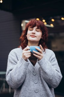 A young woman with closed eyes savors a moment with a cup of coffee. She wears a cozy gray sweater,