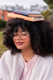 A young latin woman with curly hair and glasses balances two books on her head while outdoors in a