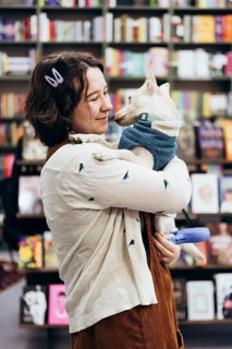 A young woman holds her dog lovingly in a warm bookstore, surrounded by colorful books. The cozy