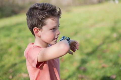 A young boy in a coral shirt examines his smartwatch in a sunny park. The background features