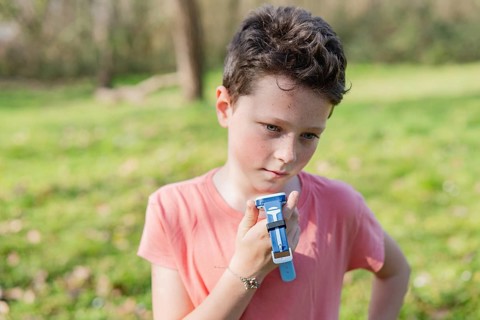 Young boy in pink shirt holding a blue smartwatch in a green park. Sunlit scene with blurred