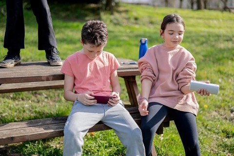 Two friends sit on a wooden bench outdoors, immersed in technology. One uses a tablet, while the
